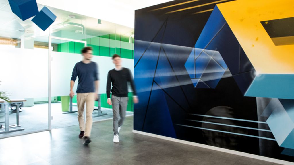 codemanufaktur_buero Zwei Mitarbeitende der codemanufaktur laufen im Büro entlang und an einem Grafftit vorbei, dass das codemanufaktur Logo abbildet.